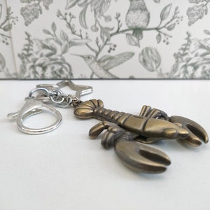 May include: A bronze-colored lobster keychain with a silver clasp and star-shaped charm. The lobster has detailed claws and body segments. The keychain is designed for attaching to keys or bags.