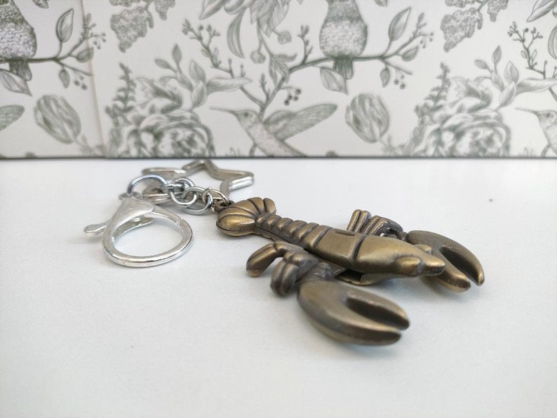 May include: A bronze-colored lobster keychain with a silver clasp and star-shaped charm. The lobster has detailed claws and body segments. The keychain is designed for attaching to keys or bags.