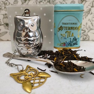May include: A silver owl-shaped tea infuser with a chain and a gold owl charm. The infuser is sitting on a spoon with loose leaf tea. A blue tin with gold lettering that reads "TRADITIONAL AFTERNOON TEA" is in the background.
