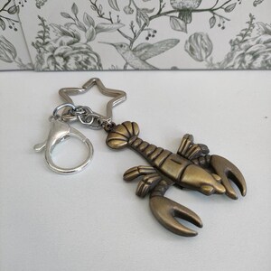May include: A bronze-colored lobster keychain with a silver star-shaped keyring and a lobster claw clasp. The lobster charm has detailed claws, body segments, and antennae. The keychain is a nautical-themed accessory.
