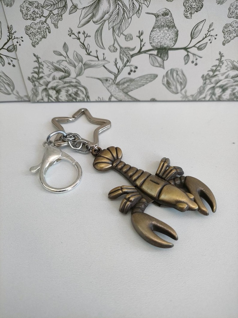 May include: A bronze-colored lobster keychain with a silver star-shaped keyring and a lobster claw clasp. The lobster charm has detailed claws, body segments, and antennae. The keychain is a nautical-themed accessory.