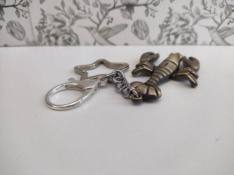 May include: A lobster-shaped keychain charm in antique brass finish. The lobster is attached to a silver-colored clasp and a star-shaped ring. The background is a white surface with a floral pattern.
