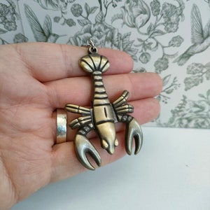 May include: A bronze-colored lobster pendant with a detailed design. The pendant features a lobster with large claws and a segmented body. It has a loop at the top for a chain. The pendant is held in a hand.