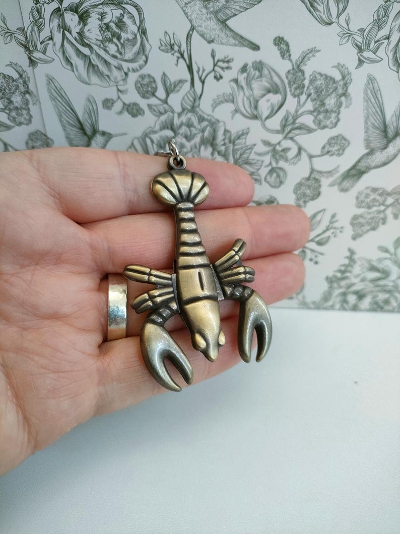 May include: A bronze-colored lobster pendant with a detailed design. The pendant features a lobster with large claws and a segmented body. It has a loop at the top for a chain. The pendant is held in a hand.