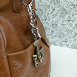 May include: A brown leather handbag with a silver-colored metal clasp and lobster charm. The bag has a front pocket and a shoulder strap. The charm is a bronze color and is attached to the bag with a chain.