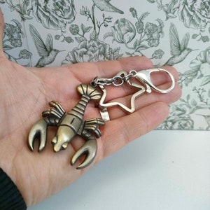 May include: A bronze-colored lobster keychain with a star-shaped charm and a silver clasp. The lobster has detailed claws and a segmented body. The keychain is held against a floral background.