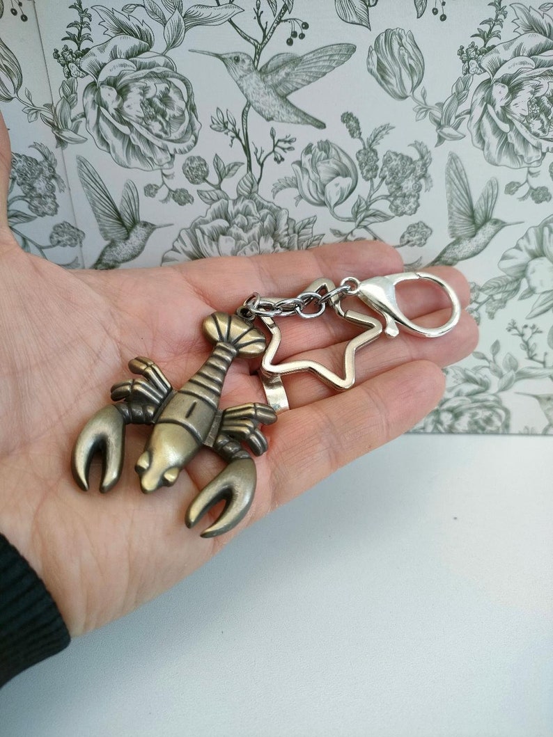May include: A bronze-colored lobster keychain with a star-shaped charm and a silver clasp. The lobster has detailed claws and a segmented body. The keychain is held against a floral background.
