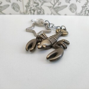 May include: A bronze-colored lobster keychain with a star-shaped charm and a silver chain. The lobster has detailed claws and a segmented body. The keychain is on a white surface with a floral patterned background.