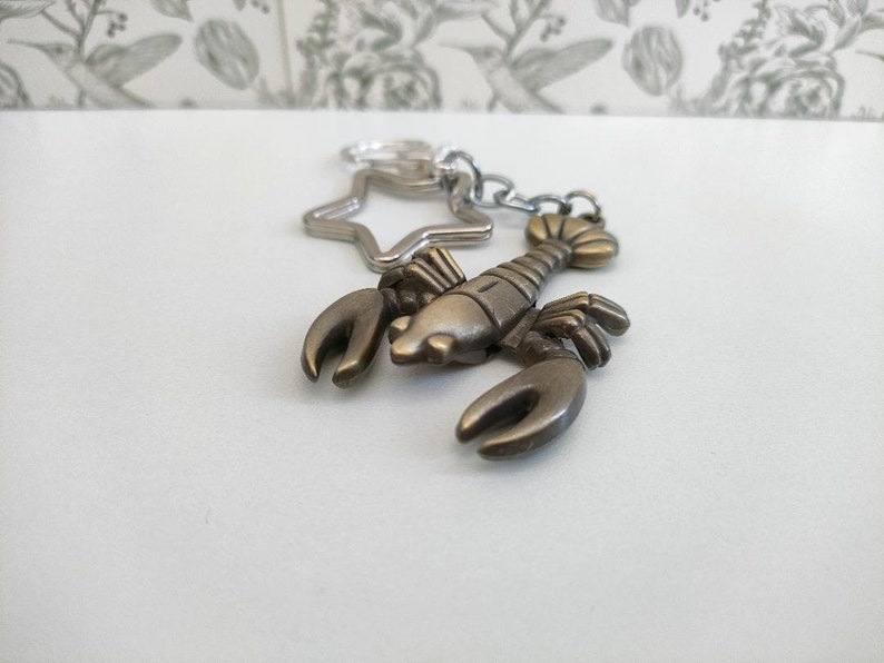 May include: A bronze-colored lobster keychain with a star-shaped charm and a silver chain. The lobster has detailed claws and a segmented body. The keychain is on a white surface with a floral patterned background.