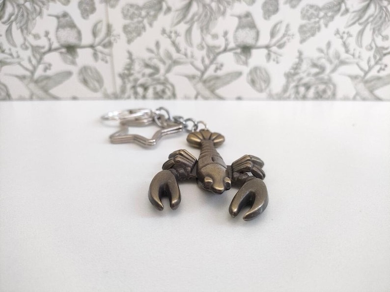May include: A bronze-colored lobster keychain with a star-shaped charm and a silver keyring. The lobster has detailed claws and a segmented body. The background is a white surface with a floral wallpaper.