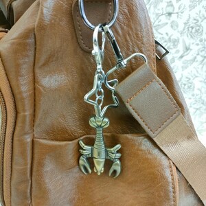 May include: A brown leather crossbody bag with a lobster-shaped charm attached to the strap. The charm is silver and bronze, and the bag has a zippered pocket and adjustable strap. The background is a floral pattern.