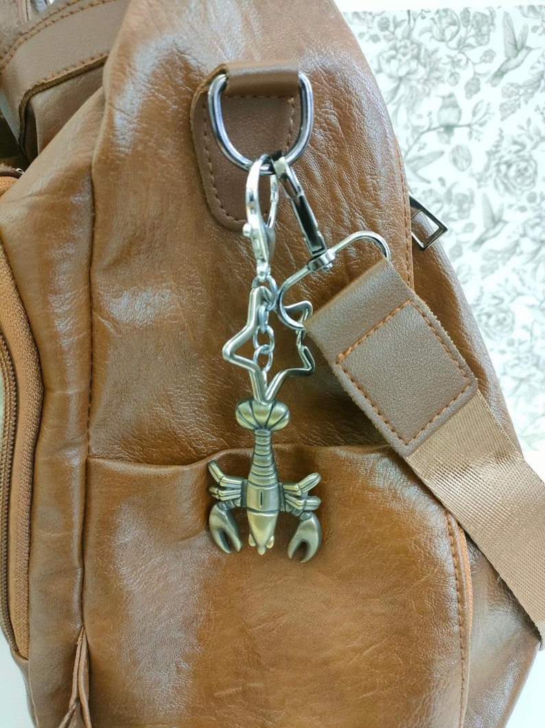 May include: A brown leather crossbody bag with a lobster-shaped charm attached to the strap. The charm is silver and bronze, and the bag has a zippered pocket and adjustable strap. The background is a floral pattern.