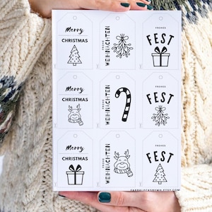 Könnte beinhalten: Ein Bogen weißer Weihnachtsetiketten mit schwarzem Text und Illustrationen. Die Etiketten zeigen Sätze wie "Merry Christmas" und "Frohe Weihnachten" sowie Bilder eines Weihnachtsbaums, eines Rentiers und eines Geschenks. Die Etiketten werden von einer Person gehalten.