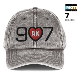 May include: A grey distressed baseball cap with a black embroidered design that says "907" with a red heart that says "AK" inside.