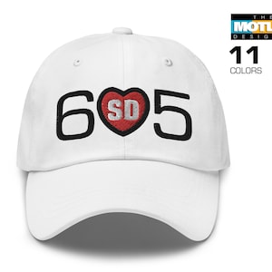 May include: White baseball cap with black embroidered text "605" and a red heart with "SD" inside. The cap has a curved brim and a white adjustable strap.