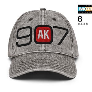 May include: A grey distressed baseball cap with black stitching and a red and black embroidered patch that says "907 AK".