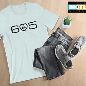 May include: A light blue t-shirt with black text that says "605" with a heart that says "SD" inside. The t-shirt is laying on a wooden surface with a pair of jeans and sneakers.