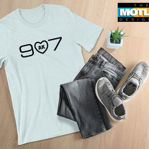 May include: A light blue t-shirt with a black graphic design that says "907 AK" with a heart in place of the "O". The shirt is laid out on a wooden surface with a pair of jeans and trainers.