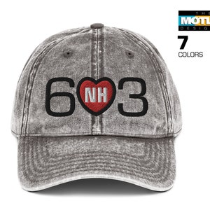 Pode incluir: Um boné de beisebol cinza desbotado com um '603' bordado em preto e um coração vermelho com 'NH' dentro.