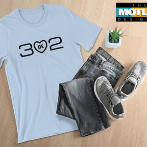 May include: A light blue t-shirt with the numbers "3", "2", and a heart symbol with "DE" inside. The shirt is paired with gray jeans and sneakers. The Motley Designer logo is in the upper right corner.