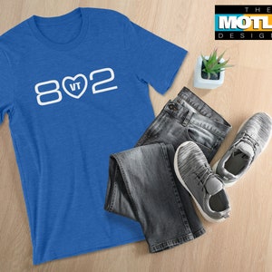 May include: A royal blue t-shirt with white text that reads "802" with a heart containing the letters "VT" in the middle. The t-shirt is laying on a wooden surface with a pair of jeans and gray sneakers.