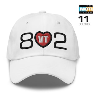 May include: White baseball cap with black embroidered text "802" and a red heart with white "VT" inside. The Motley Designer.
