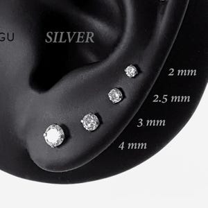 May include: Four silver stud earrings with clear round stones arranged in a row, labelled with sizes 2 mm, 2.5 mm, 3 mm, and 4 mm. The earrings are set in a dark display form, showcasing the graduated sizes of the stones from smallest to largest.