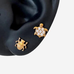 Puede incluir: Pendientes de oro. Un pendiente presenta un diseño de tortuga con piedras transparentes, y el otro un diseño de escarabajo. Los pendientes se muestran sobre una superficie negra.