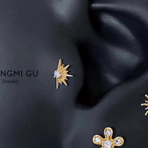 May include: A gold-toned starburst-shaped earring stud featuring a central clear gemstone, positioned on a black ear-shaped display. The text NGMI GU Jewelry is visible on the left side. The design includes radiating rays around the stone.