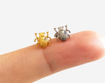 May include: Two small scarab beetle stud earrings, one gold-toned and one silver-toned, are displayed. The earrings are detailed with intricate designs and are positioned on a finger. The jewelry is from the brand Hyangmi Gu Jewelry.