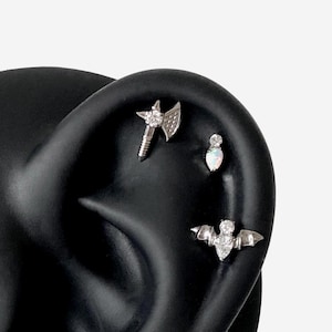 May include: Three silver-toned metal body jewelry pieces inserted into a black ear model. The top piece is an axe shape with a clear stone, the middle is a teardrop-shaped opal, and the bottom is a bat-shaped stud with clear stones.