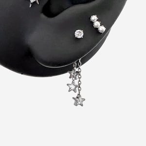 Puede incluir: Un conjunto de pendientes plateados que incluye un pendiente de piedra transparente redonda, un pendiente de tres perlas y un pendiente de cadena colgante con tres pequeños dijes en forma de estrella, cada uno con una piedra transparente.