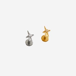 May include: Two flat back labret studs featuring a four-pointed star design. One stud is silver-toned with a clear centre stone, and the other is gold-toned with a clear centre stone. Both pieces have a circular base and a post for piercing.