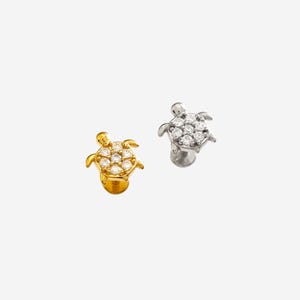 Puede incluir: Dos pendientes con forma de tortuga. Uno es dorado con piedras preciosas transparentes, el otro es plateado con piedras preciosas transparentes. Los pendientes son pequeños y detallados, con las piedras preciosas dispuestas para parecerse al caparazón de la tortuga.
