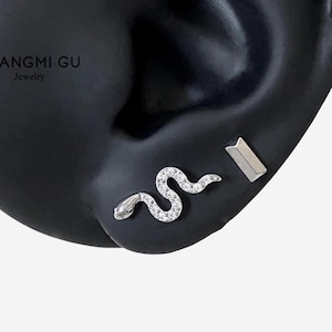 May include: A silver-toned earring set featuring a snake-shaped stud encrusted with small clear stones and a separate rectangular bar stud. The jewelry is displayed on a black ear model with the text ANGMI GU Jewelry visible in the upper corner.
