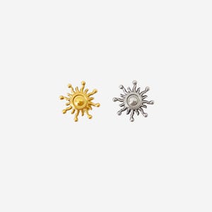 May include: Two sun-shaped metal studs, one in a polished gold tone and one in a polished silver tone. Each stud features a central circular disc surrounded by radiating, wavy rays that terminate in small, rounded spheres at the outer edges.