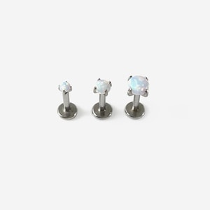 May include: Three labret studs with flat circular bases and silver-toned posts. Each stud features a prong-set, round, iridescent white opal gemstone. The studs are arranged in a row, increasing in size from left to right, showcasing a polished metal finish.