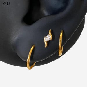 May include: A set of gold-toned jewellery consisting of two plain hoop earrings and one stud earring featuring a central clear square-cut stone set within a curved, wave-like gold metal frame, displayed on a black ear-shaped mannequin form.