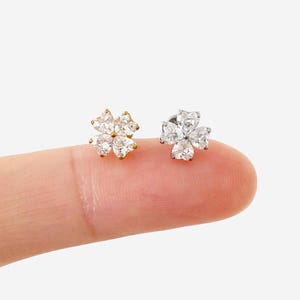 May include: Two stud earrings shaped like four-leaf clovers, each featuring four heart-shaped clear cubic zirconia stones. One earring is set in a gold-toned metal base, while the other is set in a silver-toned metal base, both displayed together.