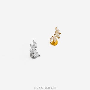 May include: Two stud earrings, one in silver-tone metal and one in gold-tone metal, each featuring a curved cluster of four marquise-cut clear stones arranged in a leaf-like pattern. The text HYANGMI GU is printed at the bottom of the image.