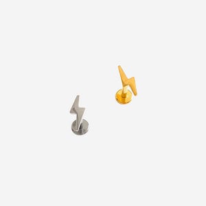 May include: Two flat back labret studs featuring lightning bolt charms. One stud is finished in a polished silver tone, while the other is finished in a polished gold tone. Each piece consists of a circular base, a post, and a lightning bolt top.