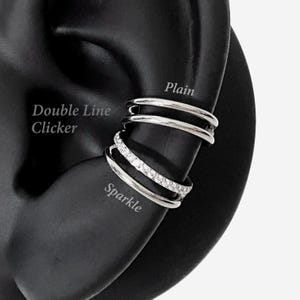 May include: A silver-toned ear cuff featuring two distinct styles: a smooth, plain double-line clicker at the top and a lower band encrusted with small, clear, sparkling stones. The text "Plain", "Double Line Clicker", and "Sparkle" are visible.