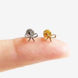 May include: Two small bow-shaped earrings, one silver and one gold, are shown. The earrings are on a finger, highlighting their size and design. The bows have a delicate, minimalist style. The earrings are likely for pierced ears.