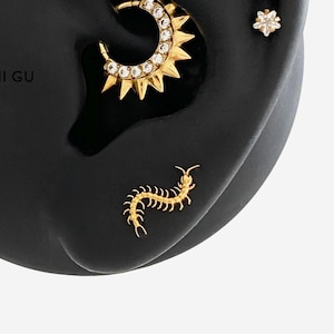 Puede incluir: Un conjunto de piercings dorados para la oreja que incluye un aro con diseño de sol y piedras transparentes, un pequeño pendiente de estrella con una piedra clara y un pendiente en forma de ciempiés sobre un modelo de oreja negro.