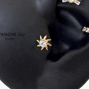 May include: A gold sun-shaped stud earring featuring a central clear round gemstone surrounded by radiating rays. The text YANGMI GU Jewelry is visible to the left. Additional small decorative stud earrings are visible in the upper right area.