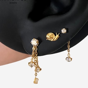 May include: A set of gold-tone earrings featuring a snail stud, a small round crystal stud, and a larger stud with three dangling gold chains tipped with clear crystals. The jewellery is arranged on an earlobe, showcasing a delicate metallic finish.