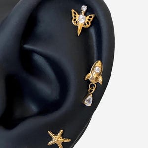 Puede incluir: Pendientes dorados con diseños celestiales y náuticos. Incluyen una estrella de mar, un cohete con una lágrima colgante y un diseño alado, todos adornados con gemas transparentes. Se muestran sobre un fondo negro.