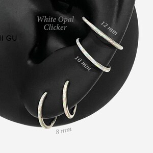 May include: Three white opal clicker hoop earrings displayed on a black ear model. The hoops are labeled with their respective diameters of 12 mm, 10 mm, and 8 mm. Each earring features a smooth, iridescent white opal inlay set into a metallic band.