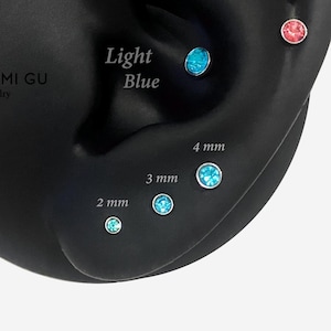 May include: A set of four round stud earrings with light blue and pink crystals set in metal. The earrings are displayed in sizes labelled 2 mm, 3 mm, and 4 mm, with the text Light Blue visible near the upper blue crystal on a black ear model.