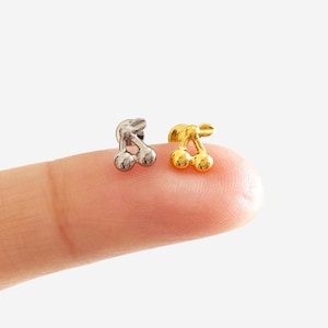 Peut inclure: Deux petites boucles d'oreilles en forme de cerise, l'une argentée et l'autre dorée, sont présentées. Les boucles d'oreilles sont posées sur un doigt, sur fond blanc. Design simple et minimaliste.